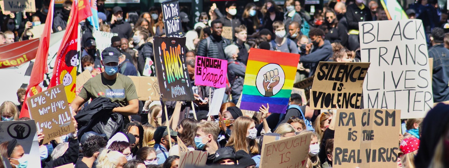 anti-racism protesters holding signs in a crowdthat say "silence is violence", "Black trans lives matter", "racism is the real virus", "Love to all who love for all", "enough is enough"